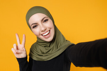 Wall Mural - Close up of excited young arabian muslim woman in hijab black green clothes doing selfie shot on mobile phone showing victory sign isolated on yellow background. People religious lifestyle concept.