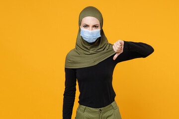 Wall Mural - Concerned young arabian muslim woman in hijab green clothes sterile face mask safe from coronavirus virus covid-19 showing thumb down isolated on yellow background. People religious lifestyle concept.