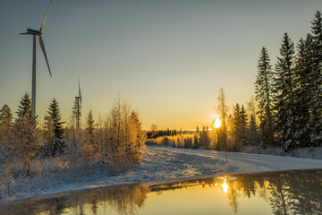 Beautiful golden winter Sunset in Scandinavian forest close to two wind turbines reflected in very clean shiny car roof. Winter country road. Sun behind forest edge