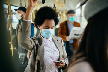 Wall Mural - African American businesswoman with face mask using smart phone while commuting by bus.