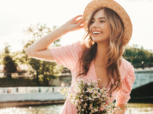 Young Beautiful Smiling Hipster Woman In Trendy Summer Sundress. Sexy Carefree Woman Posing On The Street Background In Hat At Sunset. Positive Model Outdoors On Embankment. Holding Flowers In Hands