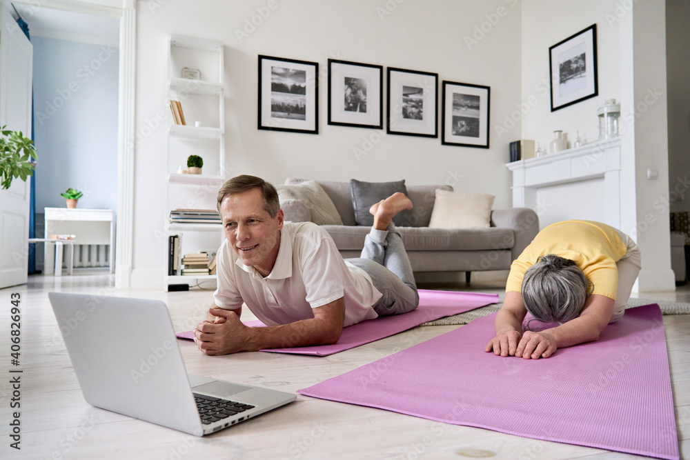 Healthy mature couple watching online live tv yoga fitness class tutorial exercising at home. Happy old middle aged husband and wife using laptop doing virtual training workout together in apartment. - obrazy, fototapety, plakaty 
