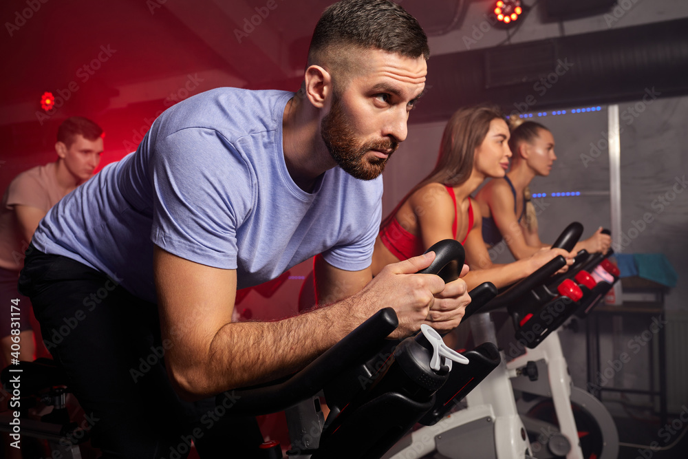 man biking in spinning class with friends at gym exercising on stationary bike, caucasian strong male is concentrated on workout - obrazy, fototapety, plakaty 