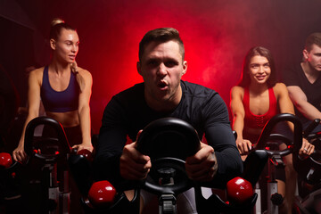 Wall Mural - man exercising cardio training on bicycle in fitness gym with friends in the background. bodybuilder, lifestyle, exercise fitness, workout and sport training concept