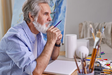 artist sitting behind table and thinking what to draw, paint. elderly male use pencil for drawings