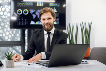 Wall Mural - Horizontal indoor shot of young handsome male office worker, sitting at the desk and smiling to camera, while working with laptop pc and making notes. Wall plasma screen on the background