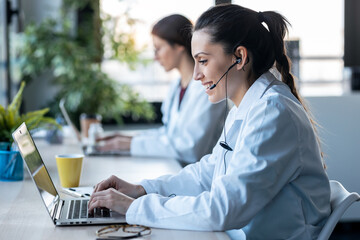 Shot of two female doctor making video call with laptop while talking with earphone sitting in the consultation.