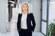 © Sofiia Shunkina - Confident blond haired businesswoman, dressed in black stylish formal suit, standing with her hands in her pockets in an office corridor in the background