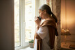 © shurkin_son - Aged people, lifestyle, retirement and loneliness. Indoor shot of elderly female with gray hair standing by window indoors, spending day at home alone, drinking hot herbal tea, having thoughtful look