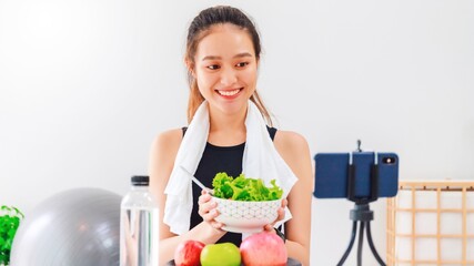 Wall Mural - Beautiful asian woman healthy blogger is showing apple fruit and clean diet food. In front of the smartphone to recording vlog video live streaming at home.Fitness influencer on social media online.