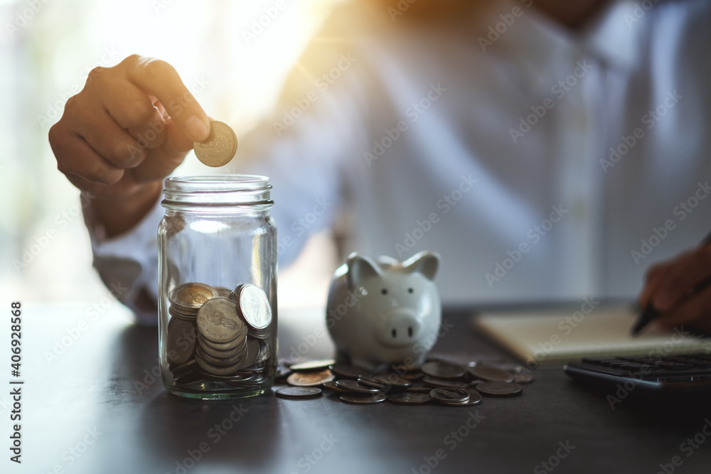 Closeup image of a woman putting coins in a glass jar and taking note with piggy bank on the table for saving money and financial concept