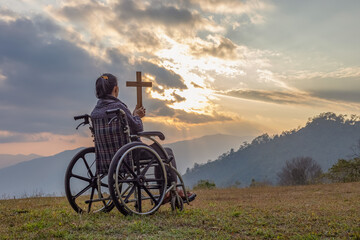 Wall Mural - Disabled handicapped woman sitting on her wheelchair and hold the crucifix in hands while praying to God at sunset background