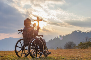 Disabled handicapped woman sitting on her wheelchair and hold the crucifix in hands while praying to God at sunset background