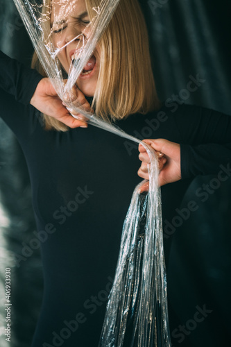 Anxiety attack. Emotional crisis. Anger violence. Art portrait of crazy disturbed psycho woman in black screaming tearing off transparent polyethylene film isolated on dark background.