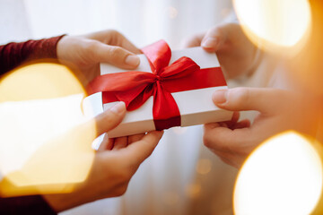 Valentine's day concept. Young romantic couple holding a gift in their hands with a garland lights bokeh on the front. White present box with a red ribbon. 14 February celebration together.