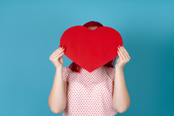 close up woman with red hair hides her face behind a large red paper heart isolated on a blue background