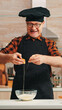 © DC Studio - Experienced baker man craking eggs for baking wearing apron enjoying hobby. Retired elderly chef with bonete mixing by hand, kneading in glass bowl pastry ingredients preparing homemade cake
