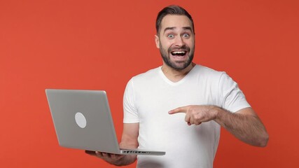 Shocked bearded young man 20s in white casual t-shirt isolated on orange color background studio. People lifestyle concept. Pointing index finger on laptop pc computer showing thumb up like gesture