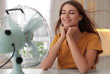 Woman Enjoying Air Flow From Fan At Table In Kitchen. Summer Heat