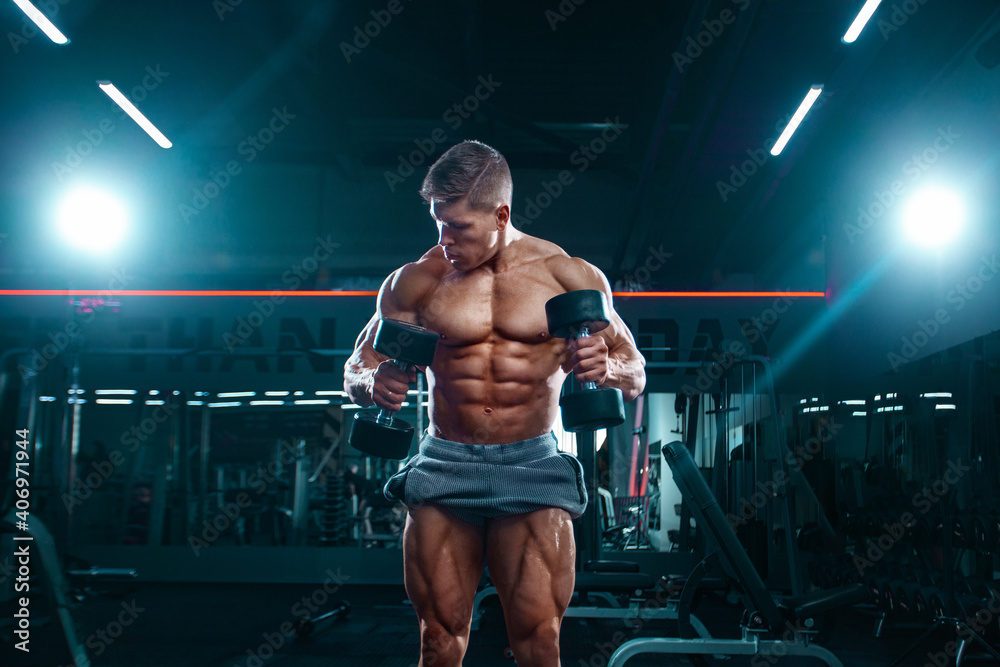 Bodybuilder athlete man pumping up muscles in the gym with dumbbells. Brutal strong muscular guy on fitness workout. Bodybuilding concept. - obrazy, fototapety, plakaty 