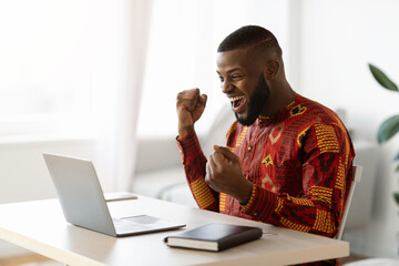Online Win. Joyful Black Man In Traditional Shirt Celebrating Success With Laptop
