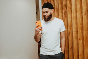 A young stylish guy, a hipster with a beautiful beard, dressed in a white, empty T-shirt with a place for your company or text, holds a smartphone and takes pictures of himself in the mirror