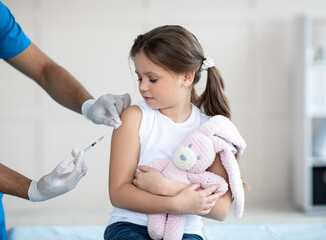 Poster - Doctor doing injection of coronavirus vaccine to little girl with toy bunny at clinic. Medicine and vaccination