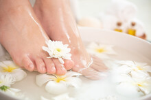 Closeup View Of Woman Soaking Her Feet In Dish With Water And Flowers On Wooden Floor. Spa Treatment And Product For Female Feet And Hand Spa. White Flowers In Ceramic Bowl.