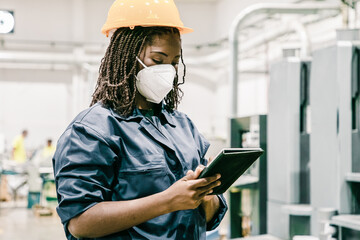 Wall Mural - African American female factory worker in mask holding tablet. Focused content skilled woman working on plant and wearing protective helmet and uniform. Manufacture and digital technology concept