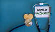 Covid-19 vaccinated symbol. White card with words 'Covid-19 vaccinated' and stethoscope on blue background. Wooden heart. Medical and covid-19 vaccinated pandemic concept, copy space.