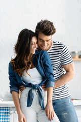 Wall Mural - happy man embracing girlfriend and smiling in kitchen