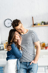 Wall Mural - man hugging cheerful girlfriend and looking away in kitchen