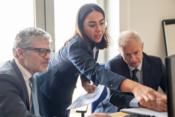 Wall Mural - Young female trade expert explaining financial charts to mature investors. Woman pointing at monitor and speaking, her colleagues listening. Medium shot. Business and expertise concept