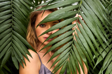 Wall Mural - woman confidently looking at camera through exotic plant leaf, serious female in nature. beauty concept