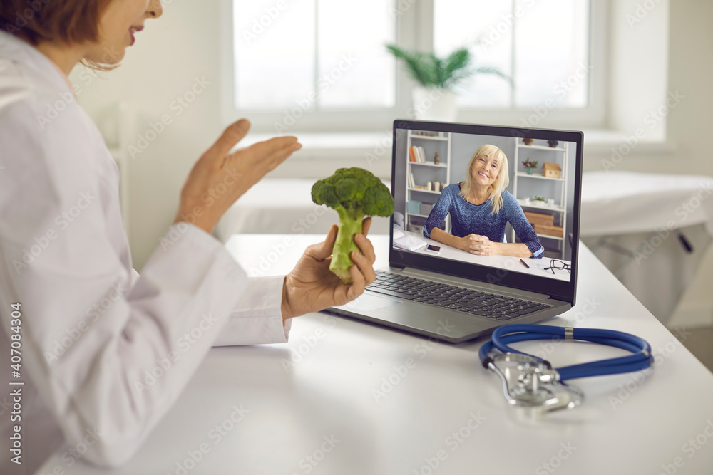 Online wellness consultation via video call: Doctor, dietitian or nutritionist sitting at laptop, holding broccoli, talking to senior patient and giving her tips on happy long life and healthy diet - obrazy, fototapety, plakaty 