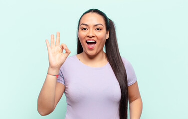 latin woman feeling successful and satisfied, smiling with mouth wide open, making okay sign with hand