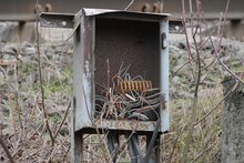 A Broken Gray Metal Box Covered In Brown Rust With Pieces Of Dangling Electrical Wires Outside