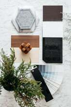Top View Of Material Selections Including Granite Tile, Marble Tile, Acoustic Tile, Walnut And Ash Wood Laminate And Painted Color Swatch With Plant And Flowers On Marble Top Table.