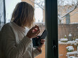 © RODRIGO - A shallow focus shot of a blonde woman with a smartphone and a mug watching snowing in front of the window