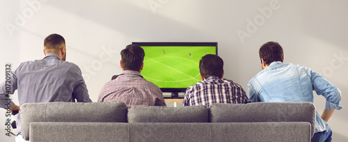 Four men friends sitting backwards on sofa at home and watching soccer footba...