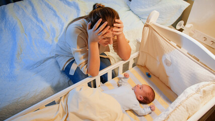 Wall Mural - Young tired mother suffering from depression lookin on her sleepless newborn baby in crib at night. Maternal depression after childbirth and sleepless nights.