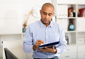 Cheerful business man in light shirt posing in office holding clipboard with papers document write notes