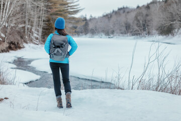 Winter sport woman walking in outdoor nature in cold snow hiking in the woods with backpack. Girl looking at frozen lake view landscape.