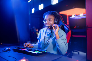 Wall Mural - Streamer african young woman rejoices in victory professional gamer playing online games computer with headphones, neon color