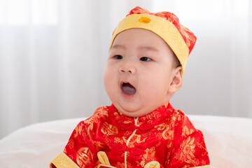 Wall Mural - Adorable Asian boy in red Chinese Costume clothes sitting on bed at home looking for somethin with curiosity, innocent cute baby feel sleepy yawning boring waiting for mother feeding