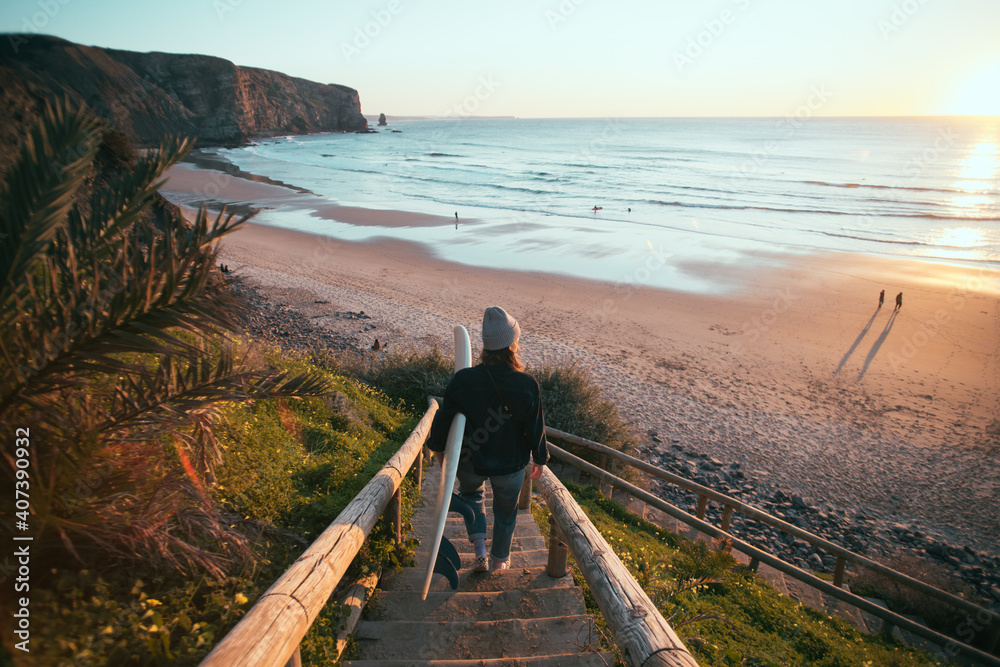 Young woman in casual clothes walk down stairs to beautiful epic beach at sunset or sunrise. Female surfer ready to start surfing lesson at dawn. Millennial lifestyle and hobby - obrazy, fototapety, plakaty 