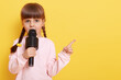 © sementsova321 - Adorable little girl with microphone on yellow background, looks at camera while talking in mic, pointing index finger aside. Copy pace for advertisement or promotional text.