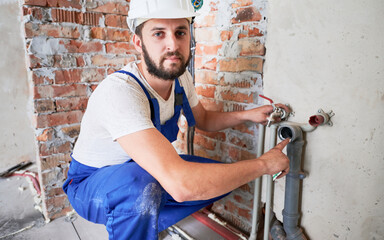 Wall Mural - Handsome male worker in safety helmet looking at camera while applying silicone grease on pipe. Man plumber in work overalls lubricating pipe while installing water system in apartment.