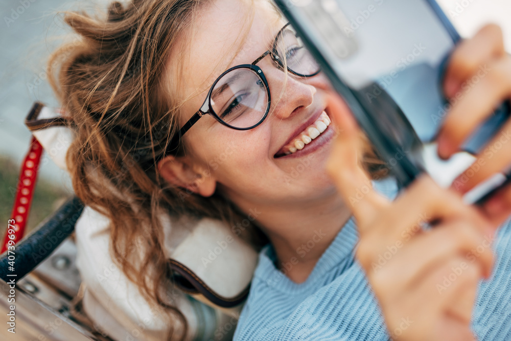 Closeup of gorgeous female in blue sweater, eyeglasses, smiling, and lying on the bench taking selfie on smart phone. Happy woman takes a rest during online conversation on a mobile phone outside.