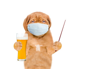 Poster - Puppy wearing medical protective face mask holds glass of beer and points away on empty space. isolated on white background